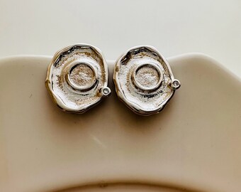 May include: Two silver-toned stud earrings with an organic, irregular shape. Each earring features a central, recessed circle with a textured surface and a small, clear accent stone. The earrings are displayed on a white surface.