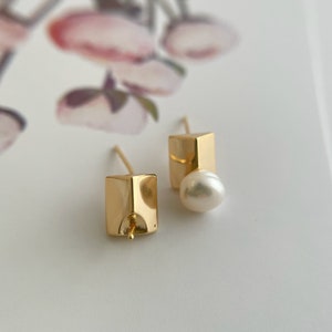 May include: A pair of gold earrings with a square shape and a pearl accent. The earrings are simple and elegant.
