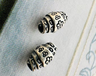 Könnte beinhalten: Zwei silberfarbene, fassförmige Perlen mit floralem Design. Die Perlen haben ein Spiralmuster mit kleinen, detaillierten Blumen und sind vor einem weichen, gemusterten Hintergrund platziert. Diese Perlen eignen sich für die Schmuckherstellung.