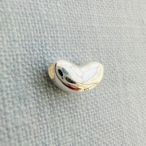 May include: A polished silver bean-shaped pendant. The smooth, reflective surface of the bean reflects the surrounding environment. The pendant has a rounded shape with a slight indentation on one side. The bean is approximately 1 inch long.