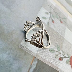 May include: A silver-colored ring with an intricate, nature-inspired design. The ring features delicate, branch-like structures with small, round accents. The design wraps around the finger, creating a unique and artistic piece of jewelry.