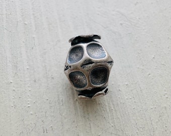 May include: A silver metal bead with a geometric design. The bead has a textured surface and is shaped like a cube with rounded edges.
