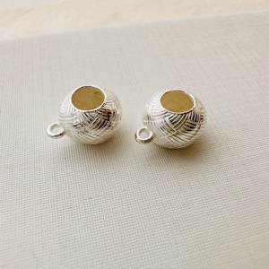 May include: Two silver bead caps with a textured design and a hole in the center. The bead caps are on a white fabric background.