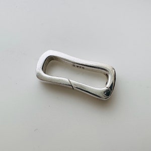 Könnte beinhalten: Ein silberfarbener Metallverschluss mit rechteckiger Form und abgerundeter Kante. Der Verschluss hat einen federbelasteten Mechanismus und ist seitlich mit "S 925" gekennzeichnet.