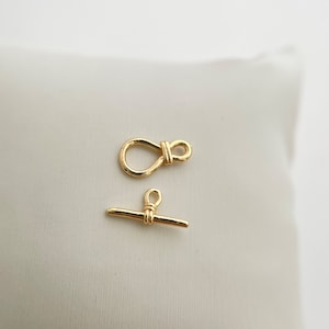 May include: Two gold-toned metal clasps for jewellery making. One clasp is a simple bar with a loop on each end. The other clasp is a loop with a knot in the centre.