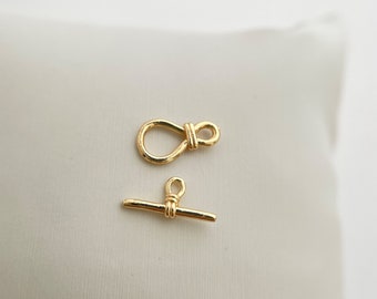 May include: Two gold-toned metal clasps for jewelry making. One clasp is a simple bar with a loop on each end. The other clasp is a loop with a knot in the center.