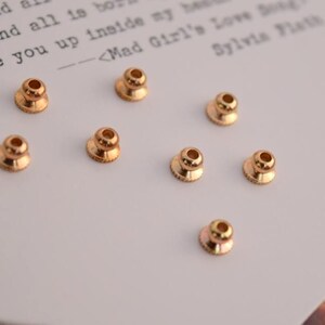 May include: Eight gold-coloured metal bead caps with a round top and a slightly flared base. The bead caps are arranged in a scattered pattern on a white surface.