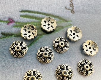 May include: A collection of antique gold-colored metal flower-shaped beads. The beads have an openwork design with a central hole and decorative swirling patterns. The beads are scattered on a light gray surface with the word "lavender" in cursive.