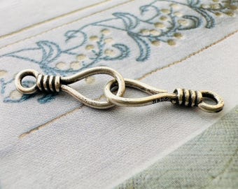 May include: A silver-colored jewelry clasp with a looped design. The clasp has a small loop on each end, with a decorative coiled wire pattern. The two loops connect in the middle. The clasp is shown against a patterned background.