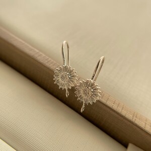May include: A pair of silver sunflower earrings with a delicate, dangling design. The earrings are made of metal and have a floral motif.