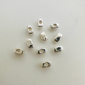 Peut inclure: Dix perles en métal argenté de forme rectangulaire avec un trou au centre. Les perles sont disposées de manière dispersée sur une surface blanche.