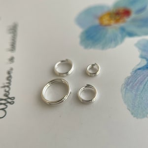 Op de afbeelding: Vier zilveren springringen in verschillende maten, gerangschikt op een wit oppervlak met een blauw bloemenontwerp op de achtergrond.