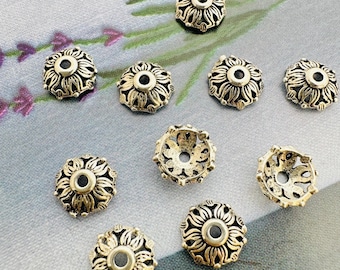 May include: A collection of silver-colored metal beads with a floral design. Each bead has a central hole and is surrounded by detailed petal-like patterns. The beads are scattered on a light gray surface with floral accents.