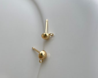 May include: A pair of gold-coloured stud earrings. Each earring features a small, spherical ball design, a post for insertion, and a tiny loop at the base. The earrings are displayed on a white surface.