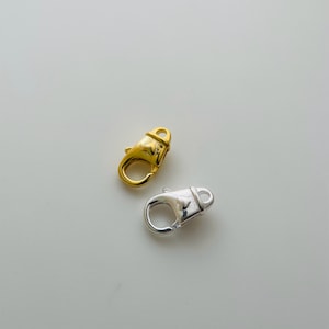 Könnte beinhalten: Zwei Hummerverschlüsse, einer in Gold und einer in Silber, werden auf weißem Hintergrund gezeigt.