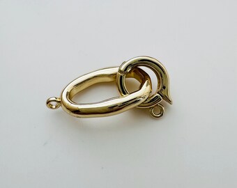 May include: A gold-toned metal clasp with a rounded oval shape and a smaller, curved hook. The clasp is designed for attaching jewelry or other items.