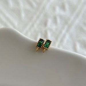 May include: A pair of gold-toned earrings with green rectangular gemstones. The earrings have a beaded border around the gemstones.