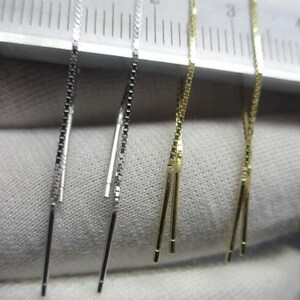 Könnte beinhalten: Zwei Paar Ohrringe, ein silbernes und ein goldenes, werden auf einem Lineal gezeigt. Die Ohrringe bestehen aus einer dünnen Kette mit einer kleinen Schlaufe am Ende.