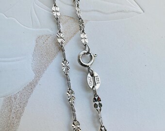 May include: A delicate silver necklace with a floral pattern. The chain is made of small, interconnected flower-shaped links. The clasp is a simple ring and hook closure. The tag reads "ITALY 925".