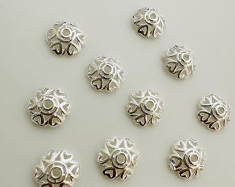 May include: A collection of ten silver-colored metal bead caps. Each cap has a flower-like design with heart-shaped cutouts around a central circular opening. The beads are scattered on a light-colored surface.