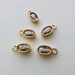 May include: Five gold-coloured oval charms with a light pink gemstone centre. Each charm has a small loop at the top for attaching to jewellery. The charms are displayed on a white surface.
