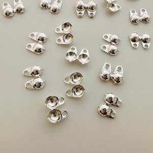 May include: A collection of small, shiny silver-coloured metal components. Each piece features a rounded, ball-like shape connected to a small, flat base with a hole. The components are scattered across a light-coloured surface.