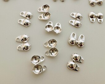 May include: A collection of small, shiny silver-colored metal components. Each piece features a rounded, ball-like shape connected to a small, flat base with a hole. The components are scattered across a light-colored surface.