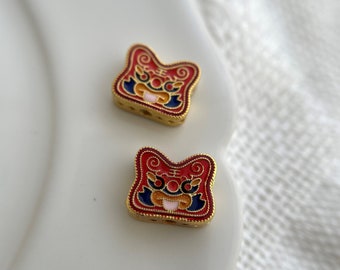 May include: Two gold-colored charms with red, blue, and white enamel designs. The charms feature a stylized lion face with intricate details.