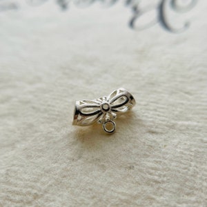 May include: Silver-toned metal jewelry finding with a decorative bow design and a small loop at the bottom.