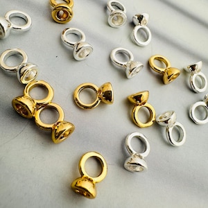 May include: A collection of gold and silver-coloured crimp end beads, used in jewellery making. Each bead has a loop for attaching to other parts. The findings are scattered on a light grey surface.