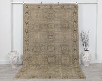 May include: A large, rectangular rug with a beige and brown geometric pattern. The rug features a repeating design of octagonal shapes with intricate detailing. The border has a decorative floral pattern. The rug is displayed against a white wall.