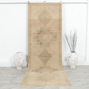 May include: A rectangular, beige-colored runner rug with a geometric pattern in shades of brown. The rug is displayed vertically, showcasing its design and texture. Two decorative vases are on either side of the rug.