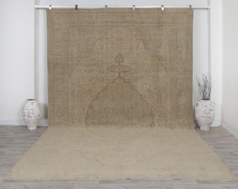 May include: A large, rectangular area rug in a muted beige tone, featuring a central mihrab-inspired arch design with intricate, subtle line patterns. The rug is displayed hanging vertically, extending onto the floor surface.
