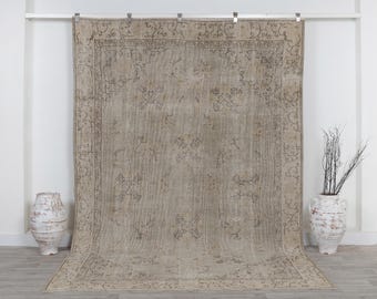 May include: A large rectangular area rug in a muted beige tone featuring a dark gray intricate floral border and symmetrical botanical motifs scattered across the center field. The rug is displayed hanging vertically against a plain wall.