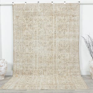 May include: A large, rectangular, beige rug with an intricate floral pattern. The rug has a distressed, vintage look with a border design. Two white decorative vases are on the left and right sides of the rug. The rug is hanging from a white bar.
