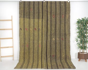 May include: A large rectangular woven rug featuring a textured olive green and brown color palette. The surface is decorated with vertical dark brown stripes and small, scattered geometric motifs in red, yellow, and brown throughout the fabric.