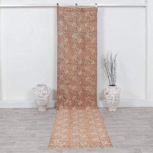 May include: A long, rectangular runner rug featuring a faded terracotta and cream-colored geometric pattern. The rug is suspended vertically from a metal rod, with its length extending down to the floor, flanked by two large, white ceramic urns.