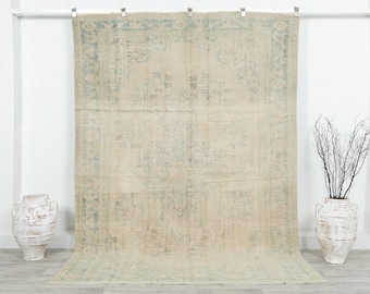 May include: A large, rectangular rug with a faded, vintage appearance. The rug features a central design with floral motifs and a border in shades of beige and light blue. The rug is hanging against a white wall, with decorative vases on either side.