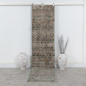 May include: A long, narrow runner rug featuring a distressed, vintage-inspired pattern in muted tones of beige, brown, and teal. The rug hangs vertically from a metal rod, displaying a repeating floral and geometric motif across its entire surface.