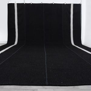 May include: A large, black rug with a white stripe running down the center and along the edges. The rug is displayed against a white backdrop, creating a striking contrast. The rug appears to be made of a thick, woven material.