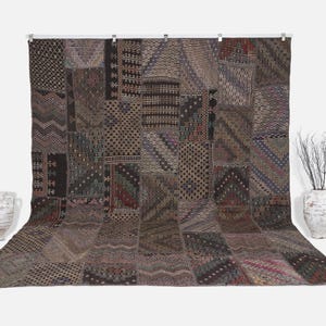 Könnte beinhalten: Ein großes Patchwork-Textil mit verschiedenen geometrischen Mustern in Braun-, Schwarz- und Grautönen. Das Textil wird vor einem weißen Hintergrund präsentiert, mit zwei weißen Vasen und einer Vase mit Zweigen auf jeder Seite.