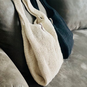 以下が含まれることがあります： グレーのソファの上に、ベージュと黒の2つの編み物トートバッグが置かれています。バッグはシンプルなステッチパターンで作られており、リラックスしたカジュアルな外観をしています。