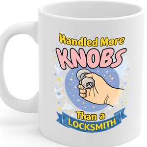 May include: White ceramic mug with a handle. The mug features the text "Handled More KNOBS Than a LOCKSMITH" in yellow, pink, and blue. A cartoon hand is holding a doorknob, set against a blue starry background.