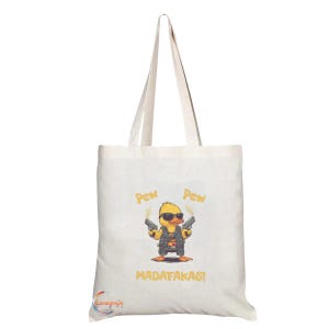 May include: Cream-coloured tote bag with a cartoon duck wearing sunglasses and a vest, holding two guns. The words "Pew Pew" are above the duck, and "MADAF*KAS!" is below. The bag has long handles.