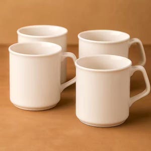 May include: Four white ceramic mugs with a simple design. Each mug has a rounded body, a sturdy handle, and a smooth, glossy finish. The mugs are arranged on a brown surface, creating a clean and minimalist aesthetic.