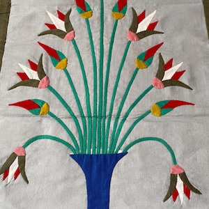 May include: A handmade textile with a blue vase holding green stems and colourful flower buds. The buds are red, white, yellow, and brown, with pink accents. The background is a light grey fabric.
