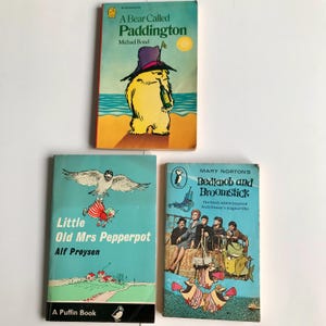 May include: Three vintage children's books. The top book is yellow with a cartoon bear on the cover and the title "A Bear Called Paddington" by Michael Bond. The middle book is blue with a cartoon bird carrying a girl on the cover and the title "Little Old Mrs Pepperpot" by Alf Prøysen. The bottom book is blue with a cartoon scene of people on a boat and the title "Bedknob and Broomstick" by Mary Norton.