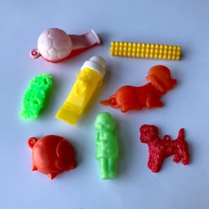 May include: A collection of nine vintage plastic toys, including a whistle, a corn on the cob, a pig, a dog, a girl, a snail, a dachshund, a green flower, and a soccer ball. The toys are all brightly colored and made of plastic.