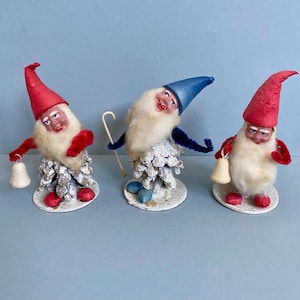 May include: Three vintage gnome figurines with red and blue hats, white beards, and bell accessories. The gnomes stand on white bases with red and blue accents. The gnomes are made of pine cones and other materials.