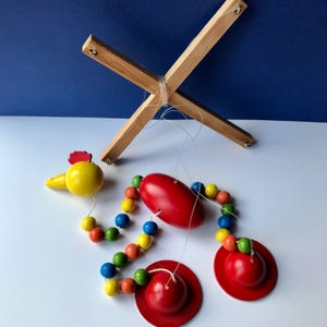 May include: A wooden baby mobile with a yellow chick, red and yellow beads, a red oval, and two red hats.